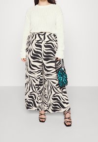 Kvinna i vit stickad tröja, svart och vit zebramönstrad vida byxor, svarta sandaler med remmar, som håller en turkos satinpåse med svart nät.