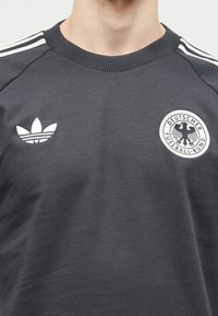 Maillot de sport gris foncé avec logo Adidas blanc sur la poitrine gauche et emblème du Deutscher Fussball-Bund sur la poitrine droite, bandes blanches aux épaules.