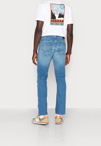 Blå denimjeans med rak passform, utrustade med bakfickor och en midjehöjd i medelhöjd. Kombinationen med en vit grafisk t-shirt och gröna sneakers.