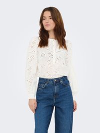 Blouse blanche en coton avec motifs floraux brodés, manches longues bouffantes et patte de boutonnage, associée à un jean bleu taille haute.