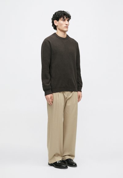 Σκούρος καφέ πουλόβερ σε υφή πλέξης, συνδυασμένο με χαλαρές μπεζ παντελόνες και μαύρα παπούτσια. Απλός σχεδιασμός με στρογγυλή λαιμόκοψη και μακριά μανίκια.