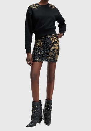 Nainen pukeutuneena mustaan paita, jossa kullanvärinen käärmeommel hartioilla, musta paljettihame, jossa kullanvärisiä käärmekuvioita, ja mustat löysät saappaat.