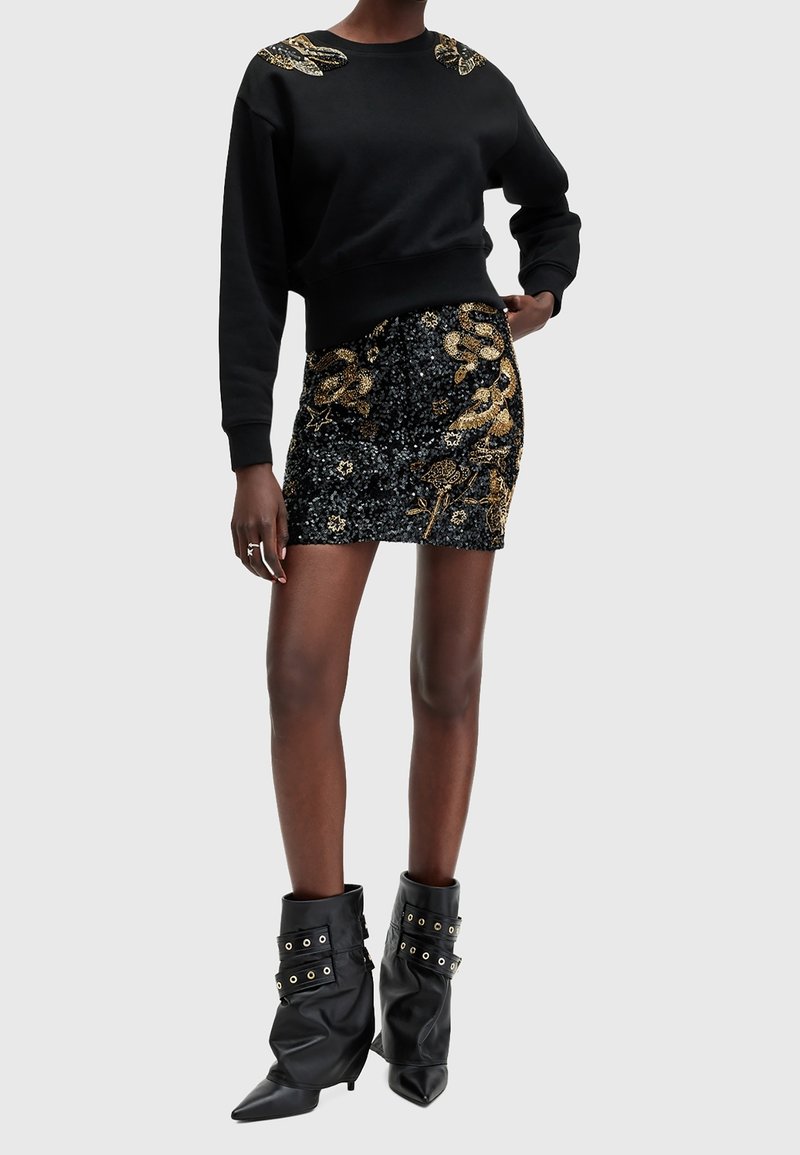 Femme portant un sweat-shirt noir avec une broderie de serpent doré sur les épaules, une mini-jupe noire à sequins avec des motifs de serpent dorés, et des bottes noires décontractées.