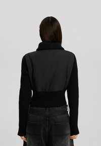 Chaqueta negra con cuello alto acanalado y espalda lisa acolchada. Mangas largas acanaladas y cintura ajustada. Punto texturizado que contrasta con la tela lisa.