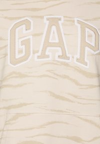 Beige ja luonnonvalkoinen huppari, jossa on suuri "GAP"-teksti tikattuna beigen sävyisinä kirjaimina, jotka on reunustettu valkoisella tigeriraidallisella kuviolla.