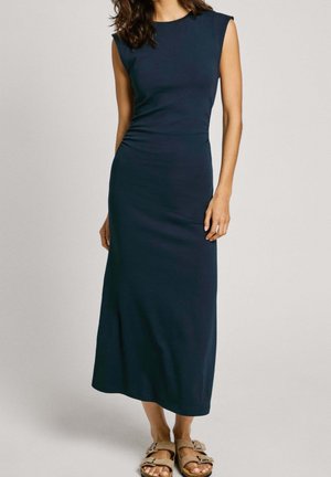 Kobieta ubrana w granatową, dopasowaną, midi sukienkę bez rękawów, z beżowymi sandałami z klamrą, stojąca na tle jasnego, jednolitego tła.