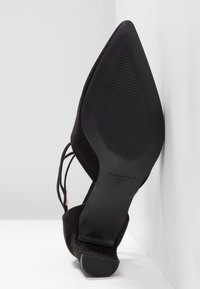 Chaussure noire en daim à talon haut avec un bout pointu et un talon fin. Dispose d'une semelle en caoutchouc texturée et de bretelles ajustables à la cheville.