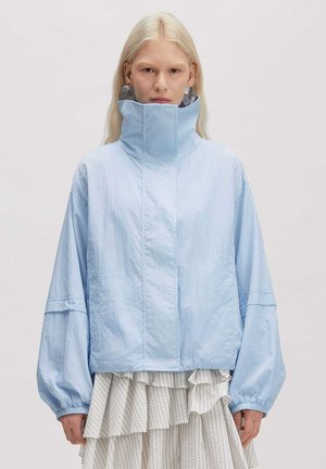 Blond person wearing oversized light blue jacket with high collar and large silver hoop earrings, paired with layered white striped skirt.