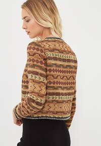 Mujer rubia vestida con una chaqueta tejida a rayas en tonos marrón, verde y burdeos, girada de lado frente a un fondo claro y sencillo.