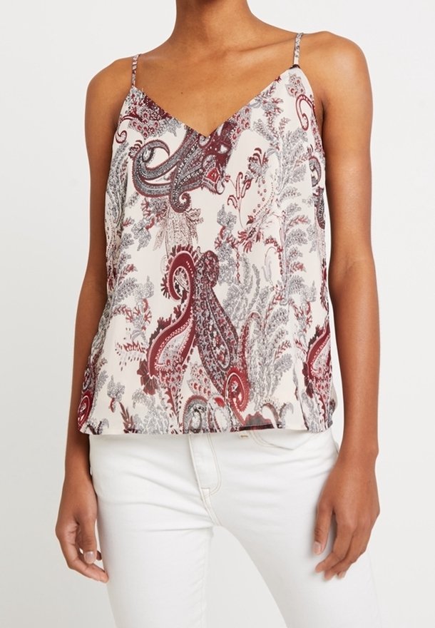 Kvinna som bär en ärmlös V-halsad linne med ett röd- och grått paisleymönster samt vita byxor mot en vit bakgrund.