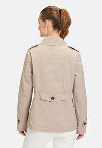 Beiger Trenchcoat mit einem strukturierten Kragen, geknöpften Schulterklappen und einem rückseitigen Knopfdetail, hergestellt aus glattem, leichtem Stoff.