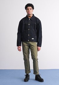 Črna denim jakna z zlatimi gumbi, v kombinaciji z olivno zelenimi hlačami. Model nosi črne škornje in stoji ob brezčasni ozadju.