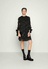 Vestido preto com tecido transparente, babados texturizados e mangas longas. Acompanhado de botas pretas até o tornozelo e meias até ao joelho. Bolsa de mão na mão.