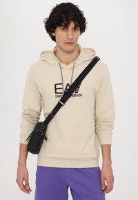 Hoodie beige con lacci e logo nero, abbinato a pantaloni corti viola. Una borsa a tracolla nera con tessuto testurizzato è indossata sulla spalla.