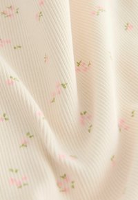 Kremasti rebrasti materijal s malim ružičastim i zelenim cvjetnim uzorkom, nježno skupljen kako bi se pokazale tekstura i detalji uzorka.