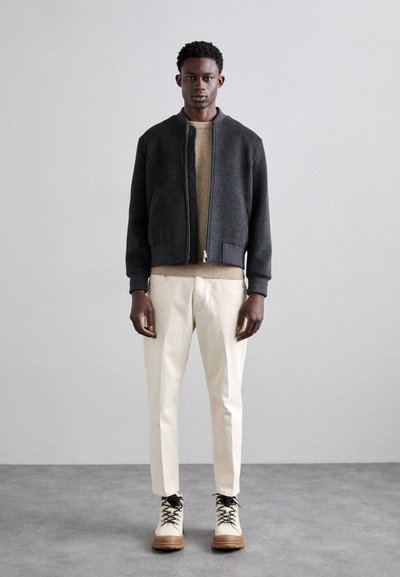 Conjunto masculino que incluye una chaqueta bomber oscura, un suéter de punto beige, pantalones crema y botas de cuero color tan con cordones negros.