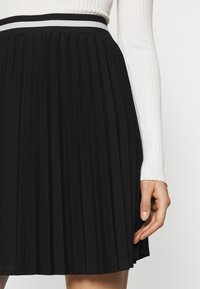 Svart plisserad kjol med en tajt midja som har en vit och grå randig detalj, med en mjuk, strukturerad textur.