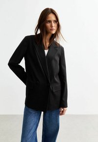 Blazer nero su misura con un fronte doppiopetto, revers a lancia e due tasche laterali, indossato sopra un top bianco e abbinato a jeans a gamba larga.