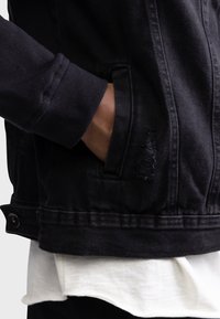 Main dans la poche avant d'une veste en jean noire portée sur une chemise blanche et une manche noire.