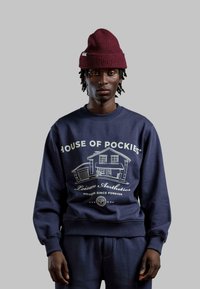 Felpa blu navy con una grafica di una casa e il testo "HOUSE OF POCKIES", con collo e polsini a coste, indossata con un berretto maroon.