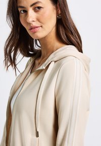 Felpa con zip beige con cappuccio regolabile, strisce laterali bicolore e una texture liscia, indossata sulla parte superiore del corpo da un modello.