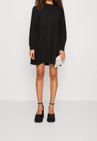 Abito lungo nero a maniche lunghe con una vestibilità rilassata, scollatura tonda e spalle arricciate, abbinato a scarpe con plateau nere e una pochette argentata.