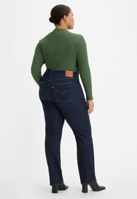 Calças de ganga de cintura alta, azul escuro, com costura laranja, bolsos traseiros e uma etiqueta de couro na cintura. Combinadas com uma blusa lista verde.