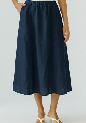 Jupe midi bleu marine avec taille élastique et poches, portée par une personne debout portant des mules blanches à talons sur un fond uni.