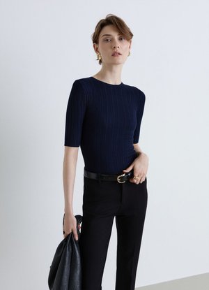 Donna in piedi davanti a uno sfondo semplice, indossa un top a coste blu navy, pantaloni neri con cintura, tiene una giacca nera e indossa orecchini dorati.