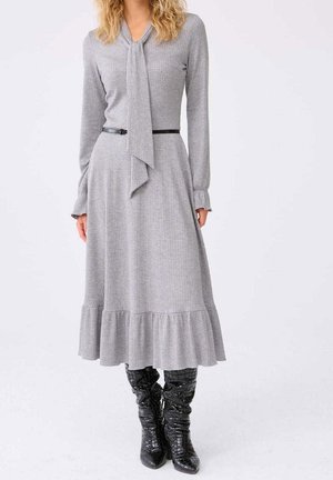 Femme portant une robe en maille grise à manches longues avec un nœud à l'avant et un ourlet à volants, assortie à des bottes noires texturées jusqu'au genou et une fine ceinture noire.