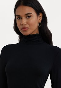 Zwarte coltrui gemaakt van een gladde, rekbare stof met lange mouwen. Beschikt over een hoge kraag en een getailleerde pasvorm. Subtiele glans.