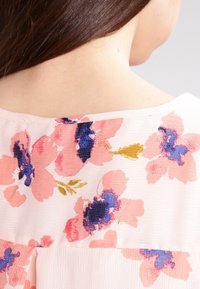 Květinová blůza z jemného růžového materiálu, se širokými růžovými a modrými květinovými vzory. Krátké rukávy a uvolněný výstřih s texturovaným detailem.