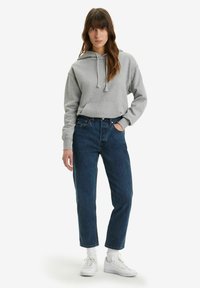 Felpa corta grigia con cappuccio regolabile, abbinata a jeans blu scuro a vita alta e sneakers bianche, indossata con calzini bianchi.