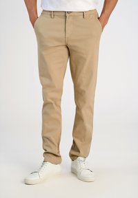 Hombre vestido con pantalones chinos beige, con las manos en los bolsillos y zapatillas blancas, de pie frente a un fondo claro y liso.