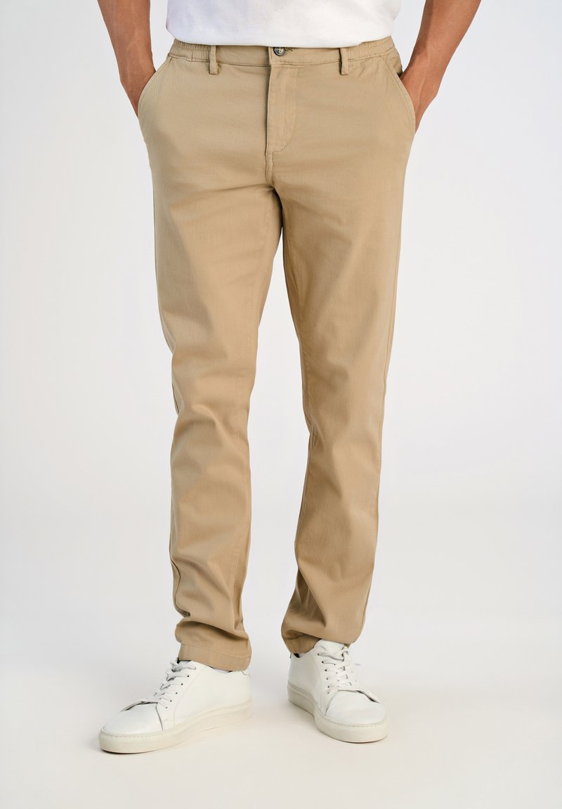Hombre vestido con pantalones chinos beige, con las manos en los bolsillos y zapatillas blancas, de pie frente a un fondo claro y liso.