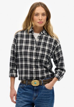 Camisa de cuadros en blanco y negro con cuello abotonado, con mangas arremangadas. Combinada con un cinturón marrón y jeans azules.
