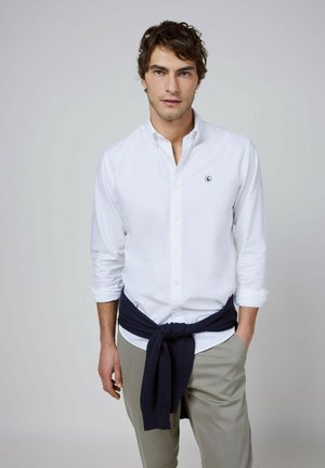 Hombre con camisa blanca abotonada con las mangas remangadas, pantalones beige y un suéter oscuro atado alrededor de su cintura sobre un fondo sencillo.