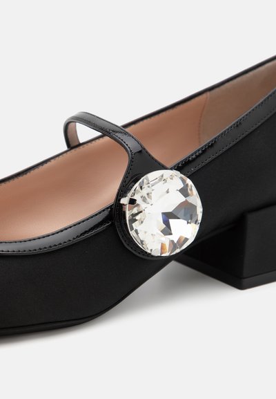 Μαύρες πατινέ δερμάτινες Mary Jane με χαρακτηριστική μεγάλη διάφανη πέτρα στο πλάι και χοντρό τετράγωνο τακούνι. Λείος υφής.
