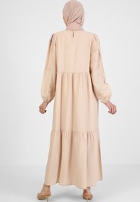 Dlouhé, splývavé šaty v broskvové barvě vyrobené z jemného materiálu. Mají dlouhé rukávy, vrstevnatý design a květinovou výšivku na ramenech.