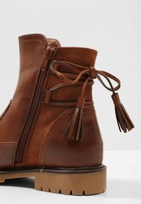 Bottines en cuir marron avec un bout rond, une fermeture éclair sur le côté et un lien décoratif avec des pompons à l'arrière. Surface texturée et semelle épaisse.