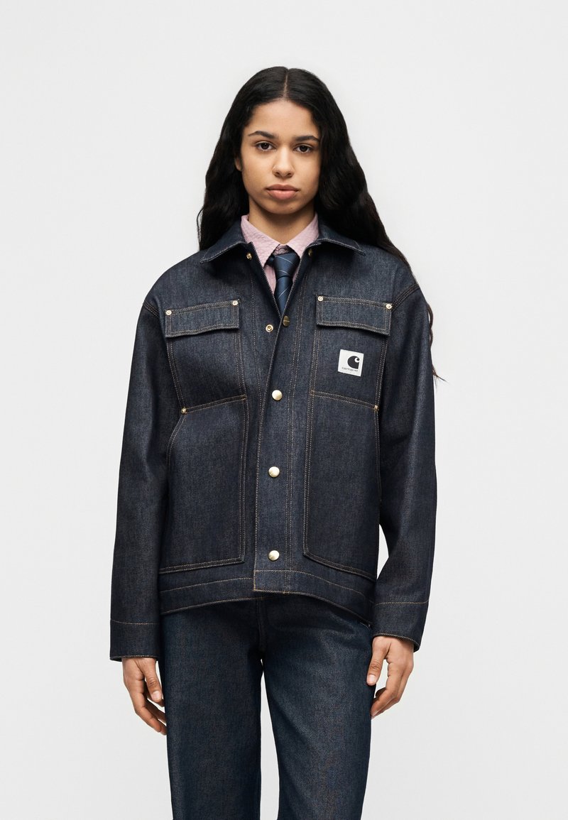 Veste en denim foncé avec boutons dorés, deux poches poitrine et un patch logo. Elle présente un col et une coupe droite, associée à un pantalon en denim assorti.