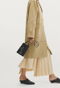 Beige ruskinds lang frakke med to knapper, kombineret med en plisseret cremefarvet nederdel og sorte lædermules. Sort håndtaske med tynde stropper i hånden.