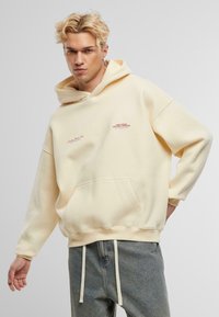 Kremowa, oversize bluza z kapturem z miękkiego materiału, z frontową kieszenią, sznurkiem do regulacji kaptura oraz czerwonymi akcentami tekstowymi na klatce piersiowej.
