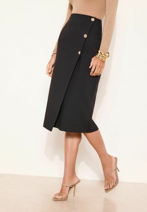 Femeie purtând o fustă neagră tip petrecută cu trei nasturi decorativi, top bej cu mâneci lungi, brățări aurii și sandale bej cu toc înalt.