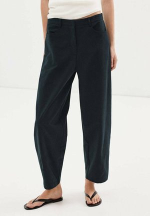 Personne portant un pantalon noir ample et des sandales noires en tongs, debout sur un sol blanc devant un fond uni.