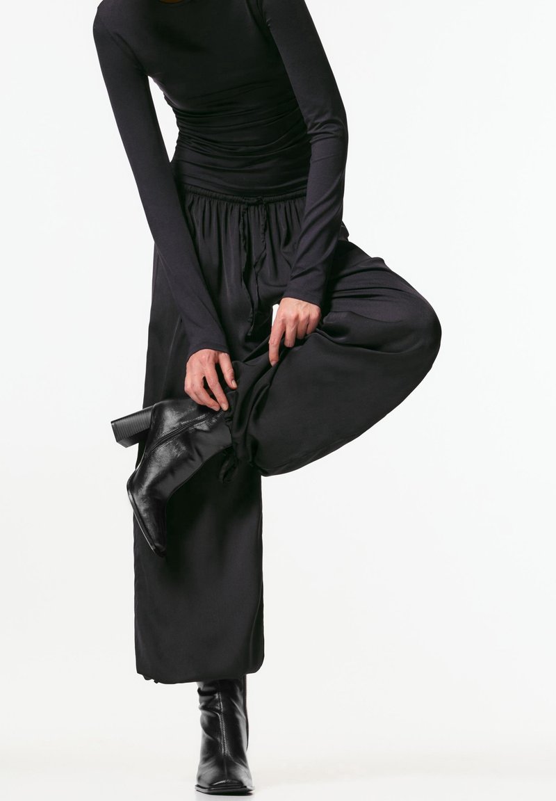 Personne portant un haut noir à manches longues, un pantalon noir ample et des bottines noires à talons, ajustant la jambe du pantalon en équilibrant sur un pied.