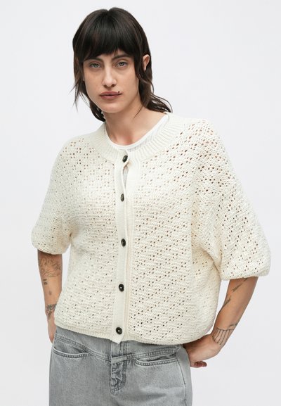 Κρεμ πλεκτή ζακέτα με κοντά, φουσκωτά μανίκια, ανοιχτό πλεκτό μοτίβο, κούμπωμα στο εμπρός μέρος με μαύρα κουμπιά. Συνδυάζεται με γκρι παντελόνι.