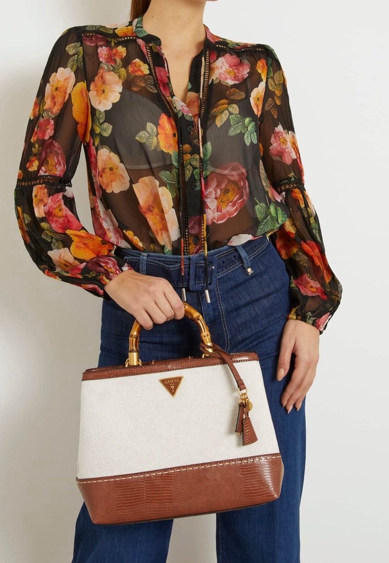 Femme en blouse noire transparente avec imprimé floral vif et jean bleu tenant un sac à main beige et marron avec des poignées en bambou.
