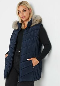 Laivastonsininen puffertakki vetskari edessä, vaakasuora tikkauskuvio, turkisreunustettu huppu ja sivutaskut, päällä musta pitkähihainen paita.