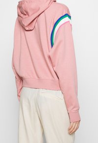 Sudadera corta rosa hecha de material suave, con rayas multicolores (azul, verde, rosa, blanco) a lo largo de los hombros y puños acanalados.